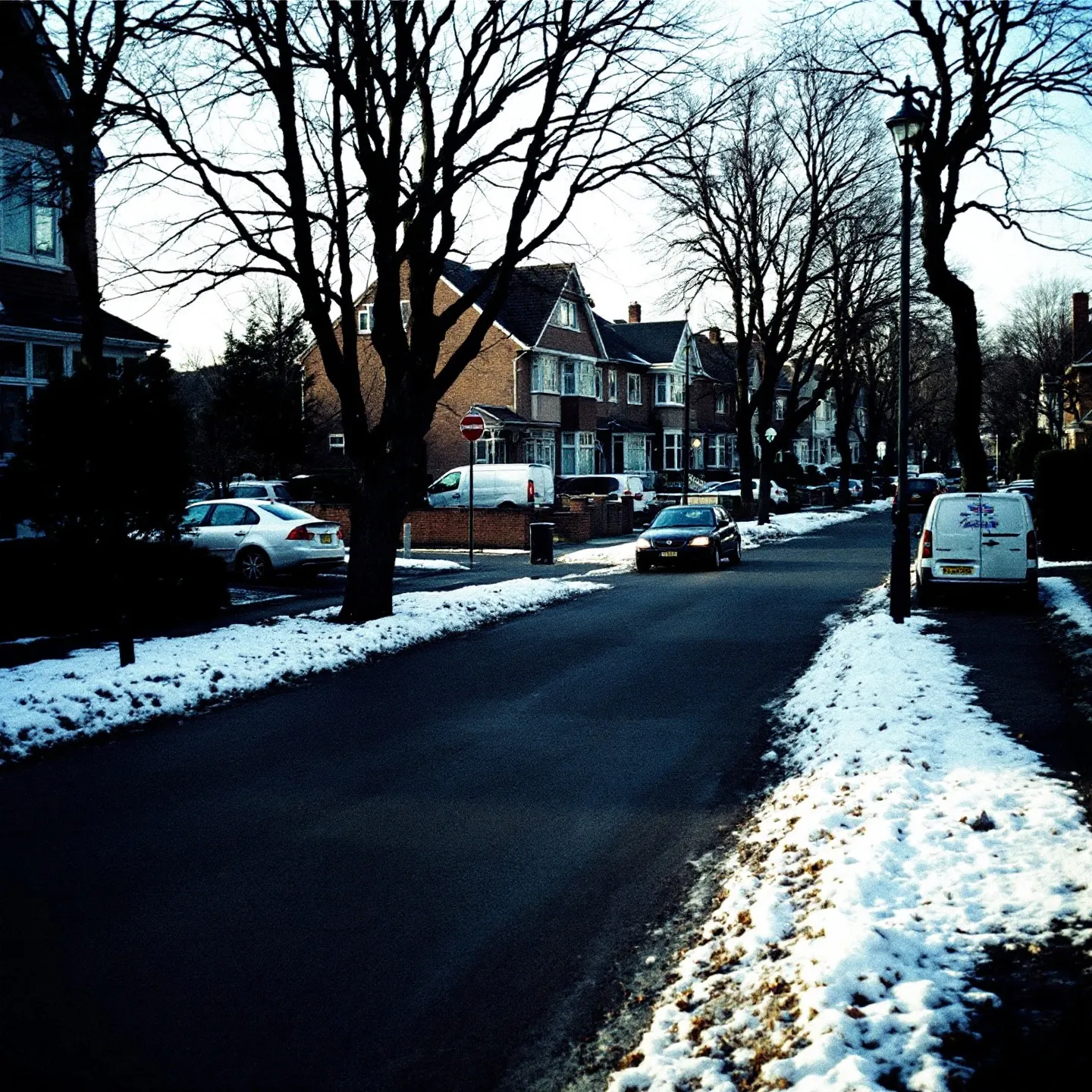 The same kind of suburban street in winter, bare trees and old snow at the kerb.